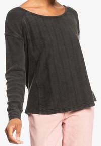 Vrouw in een donkergrijze top met lange mouwen en een textuur met verticale strepen, gecombineerd met lichtroze broek, staande tegen een witte achtergrond.