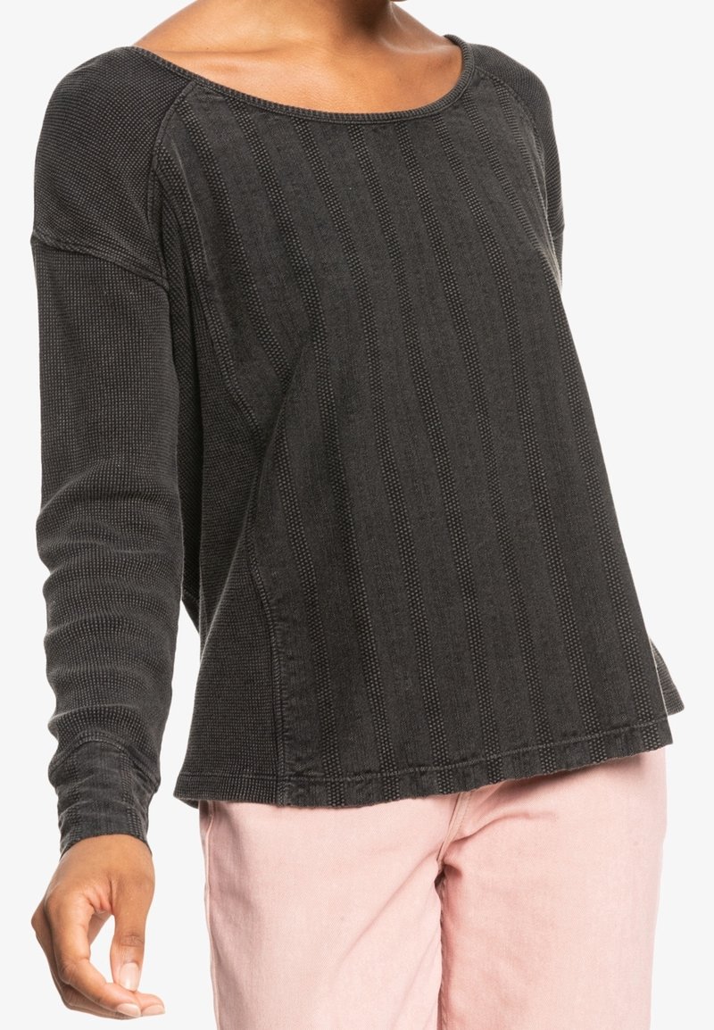 Vrouw in een donkergrijze top met lange mouwen en een textuur met verticale strepen, gecombineerd met lichtroze broek, staande tegen een witte achtergrond.