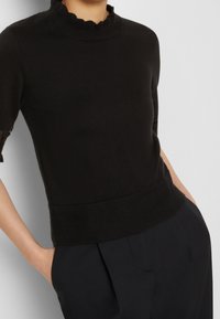 Top de punto negro con cuello falso festoneado, mangas cortas y dobladillo acanalado. Presenta una textura suave y un diseño ajustado.