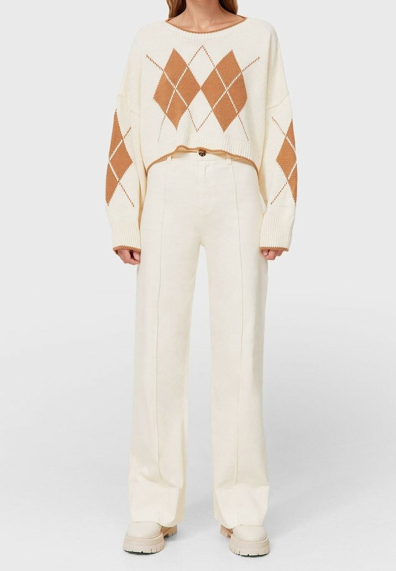 Femme portant un pantalon large taille haute crème associé à un pull court blanc avec des motifs losanges bruns sur le devant et les manches.