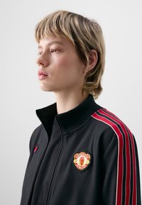 Črna jakna z zadrgo, ki ima rdeče črte na rokavih in pečat Manchester Uniteda na prsih. Mehak, teksturiran material z visokim ovratnikom.