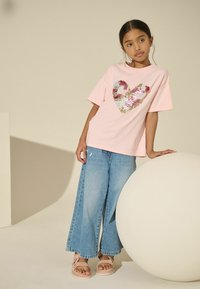 Helles pinkes T-Shirt mit einem großen, mehrfarbigen Paillettenherzdesign. Kombiniert mit blauen weiten Jeans und beigen Sandalen. Glatte Textur.