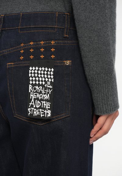 Jeans in denim scuro con cuciture arancioni visibili, dotati di un'etichetta nera unica che mostra del testo bianco e un motivo a croce.