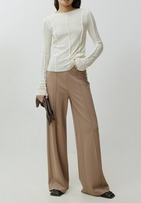 Langearmige, off-white top met textiel structuur en frontknopen, gecombineerd met hooggewaisted, wijde beige broeken en zwarte handschoenen.