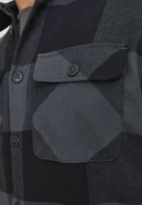 Zwart en grijs flanellen overhemd met een geruit patroon. Heeft twee borstzakken met zwarte knopen en een gestructureerde stof.