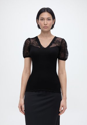 Žena s krátkymi čiernymi vlasmi, oblečená v čiernom topu s priesvitnými čipkovými nafúknutými rukávmi a výstrihom do V, stojaca pred jednoduchým bielym pozadím.