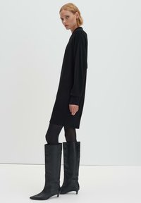 Abito nero a maniche lunghe dal taglio morbido, con spacchi laterali, indossato con stivali neri in pelle al ginocchio con tacco. Sfondo semplice e testurizzato.