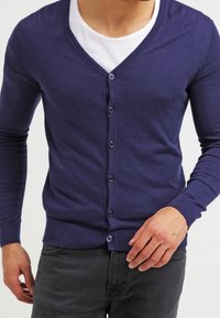 Mörkblå cardigan i mjukt material, med V-ringning, långa ärmar och knäppning, bärs över en vit T-shirt.