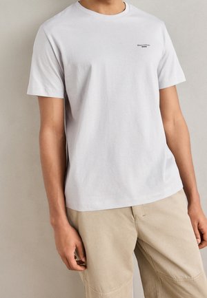 Mężczyzna ubrany w prostą białą koszulkę z krótkim rękawem z małym czarnym logo "Marc O'Polo Denim" na piersi, beżowe spodnie, stojący na neutralnym tle.