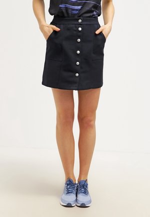 Vrouw draagt een zwarte rok met knoopsluiting en zakken, een donker patroon top en lichtblauwe sneakers, staande met handen in de zakken.