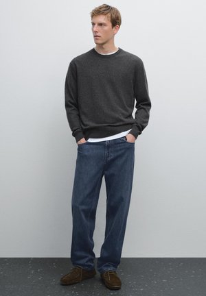 Grauer Strickpullover mit Rundhalsausschnitt, über einem weißen Hemd getragen, kombiniert mit lockeren blauen Jeans und braunen Wildlederschuhen.