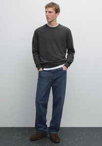 Szary sweter dzianinowy, biały podkoszulek z okrągłym dekoltem na dole, luźne niebieskie dżinsy, brązowe zamszowe buty, stojący na jasnym tle.