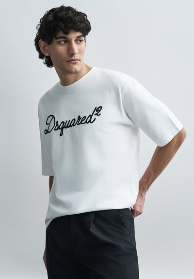 Λευκό βαμβακερό T-shirt με κοντά μανίκια και στρογγυλή λαιμόκοψη, με μαύρο κεντητό λογότυπο "Dsquared²" στο μπροστινό μέρος. Συνδυάζεται με σκούρο παντελόνι.