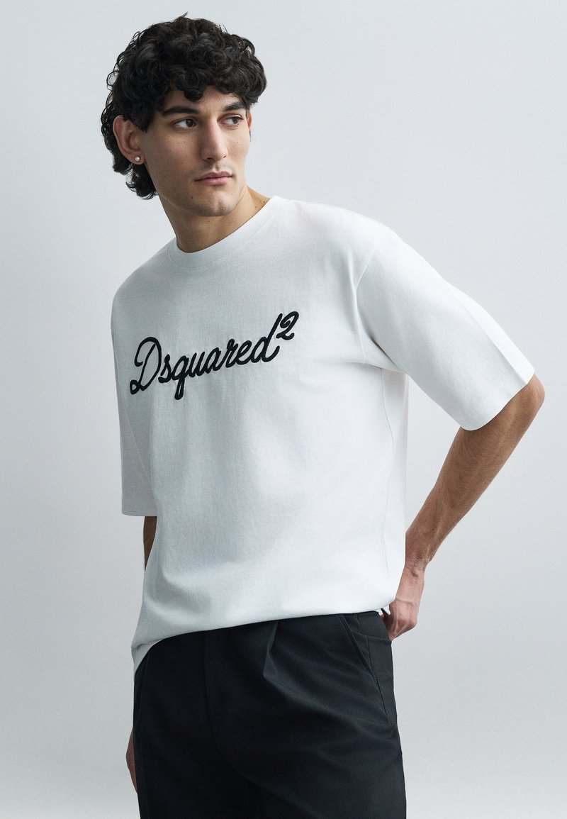 White cotton T-shirt with short sleeves and a crew neck, featuring black embroidered "Dsquared²" logo on the front. Paired with dark pants.