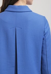 Blue textured blazer with a notched collar, featuring a back pleat and clean lines, made from a lightweight, structured fabric.