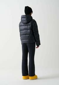 Mustmustori jakk, millel on kapuuts, tikitud disain, koos tumedate pükste ja kollaste jalatsitega. Korrusel on sile tekstuur ja lihtsad jooned.