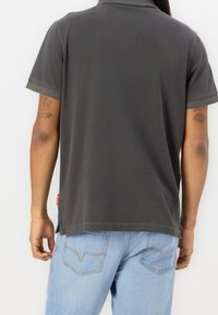 Personne portant une chemise gris foncé à manches courtes et un jean bleu clair, vue de dos avec les bras tatoués visibles.