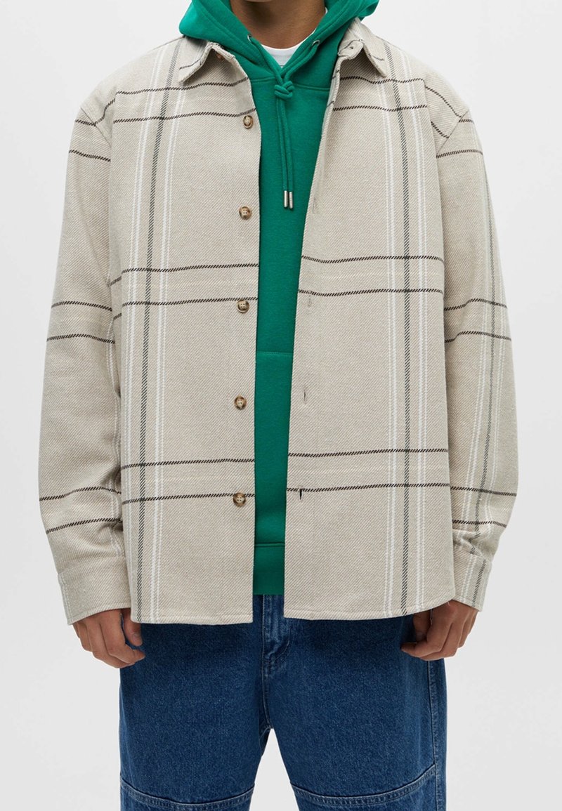 Beige geruite shirtjack gemaakt van textuurstof met bruine en witte strepen, met knoopsluitingen en een losse pasvorm over een groene hoodie.