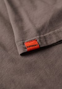 Camiseta de algodón gris con una pequeña etiqueta tejida naranja con el texto "SUPERDRY" en el dobladillo, que muestra una textura suave y un diseño simple.