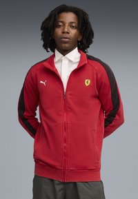 Rød zip-up jakke med sorte sidepaneler, prydet med Puma-logo og Ferrari-emblem på brystet. Blødt stof med ribbede manchetter og kant.