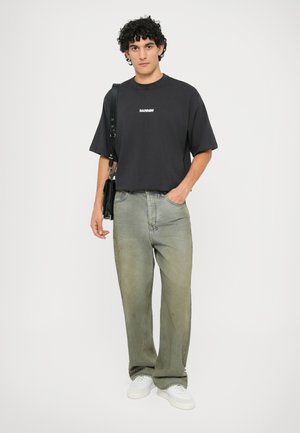 Black cotton t-shirt with white logo, paired with light green baggy denim jeans, and white sneakers. Model holds a black shoulder bag.
