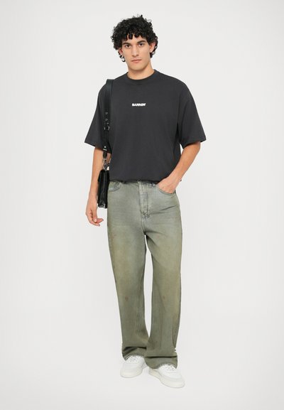 T-shirt en coton noir avec logo blanc, associé à un jean baggy en denim vert clair et des baskets blanches. Le modèle tient un sac à bandoulière noir.
