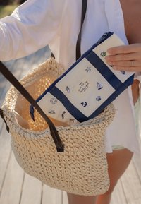 Main tenant une pochette bleue et blanche avec des motifs nautiques, la plaçant dans un sac en paille tressée avec des poignées foncées.