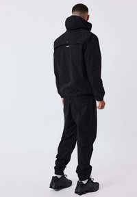 Uomo con giacca nera con cappuccio e pantaloni neri abbinati, con sneakers nere, in piedi e rivolto di spalle su uno sfondo chiaro e uniforme.