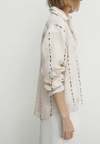 Camicia di lino chiaro con maniche arrotolate, caratterizzata da motivi di ricamo lineare neri. Collo classico e design dalla vestibilità rilassata.