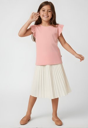 Uśmiechnięta dziewczyna w różowej bluzce na ramiączkach z falbankami i białej plisowanej spódnicy pozuje z jedną ręką przy uchu, a drugą wyciągniętą.