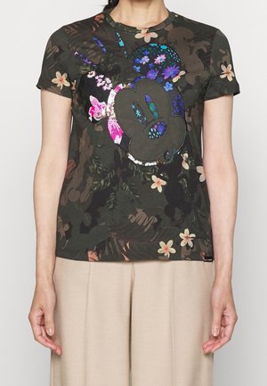 Frau trägt ein dunkles Blumen-T-Shirt mit einem großen, bunten Mickey-Mouse-Gesicht als Design, kombiniert mit hellbeigen Hosen.