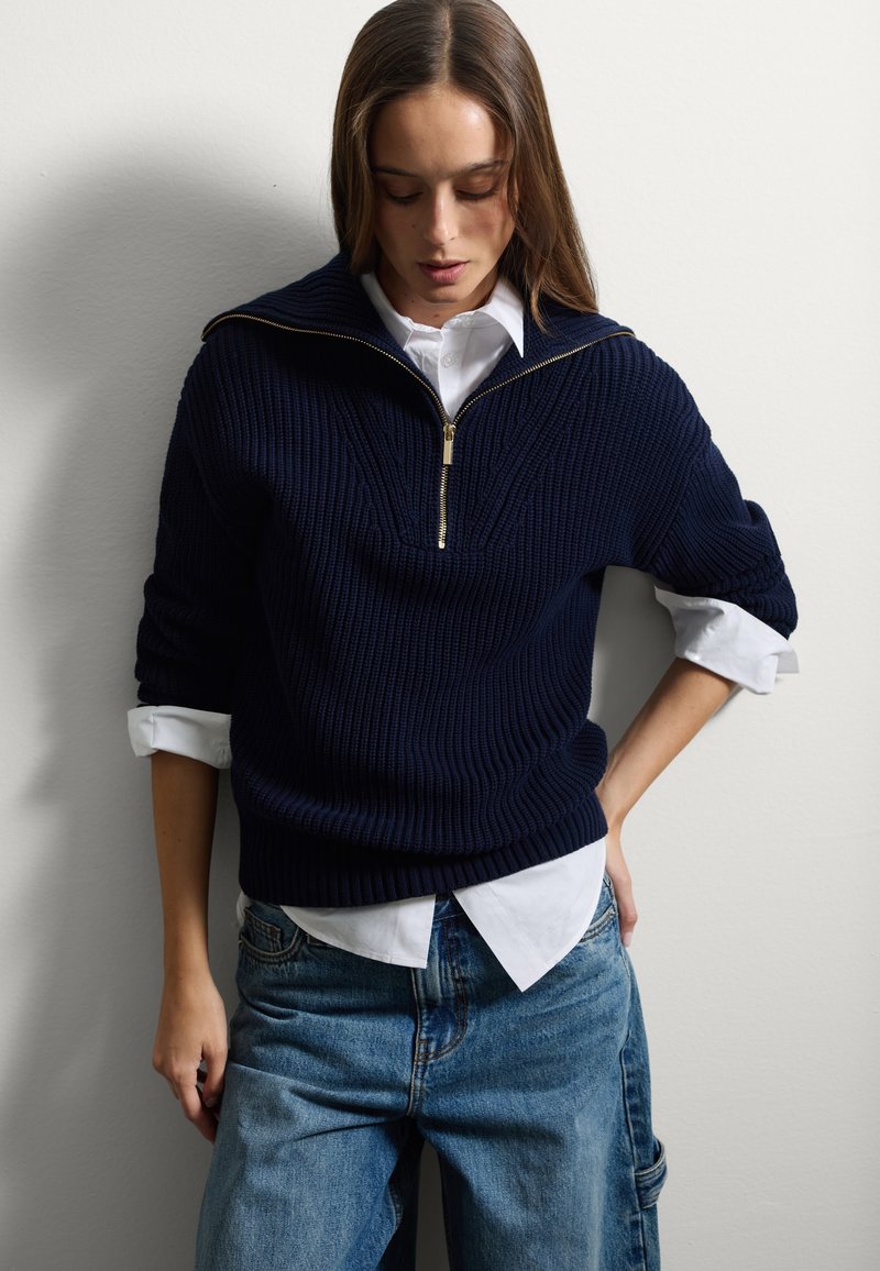 Pull en tricot côtelé marine avec un col zippé mi-haut et une chemise blanche en dessous. Le pull a une coupe ample et des poignets à manches roulées.