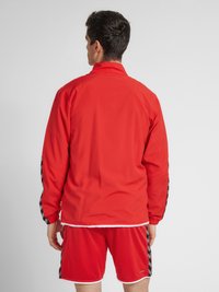 Rød atletisk jakke med høj krave og elastiske manchetter, der har sorte og grå mønstrede detaljer langs ærmerne. Bæres med matchende shorts.