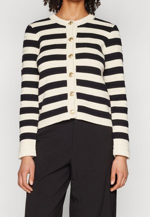Frau trägt eine schwarz-weiß horizontal gestreifte, geknöpfte Strickjacke mit goldenen Knöpfen und schwarze Hose, steht vor einem schlichten Hintergrund.