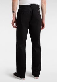 Uomo che indossa pantaloni dritti neri e sneakers bianche e nere, in piedi con la schiena rivolta verso la fotocamera su uno sfondo bianco semplice.
