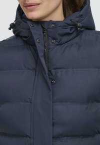 Gros plan du visage d'une femme, portant une doudoune bleu marine avec un col montant et une capuche ajustable, mettant en avant une fermeture éclair et des boutons-pression.