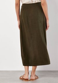 Frau, die einen knöchellangen, dunkeloliven Rock und sandfarbene Ballerinas trägt, steht auf einem glatten grauen Boden vor einer weißen Wand.