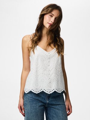 Giovane donna con capelli castani ondulati che indossa un top senza maniche in pizzo occhiellato bianco e jeans blu su sfondo bianco uniforme.