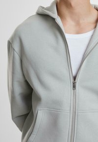 Hellgraue Zip-Hoodie aus weichem Stoff, mit Kapuze, Fronttaschen und einem glatten Reißverschluss. Das Innere wird durch ein weißes Oberteil darunter ergänzt.