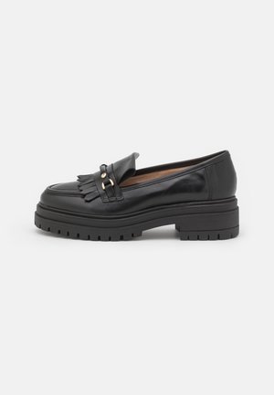 Mocassin en cuir noir avec semelle crantée épaisse, détail à franges et boucle dorée sur un fond blanc, vue de côté.