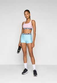 Sport-BH in zartem Pink mit schwarzem "UNDER ARMOUR"-Bund; passende hellblaue Shorts. Stehend, hält eine schwarze Wasserflasche, trägt schwarze Sneakers.