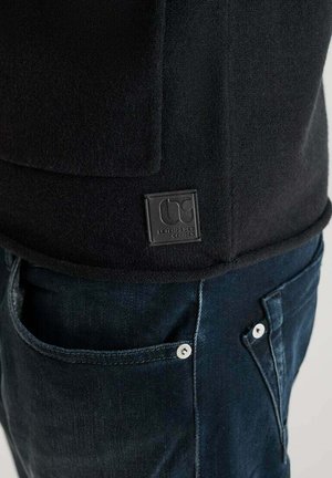 Primo piano dell'orlo di una giacca nera con texture, con una piccola etichetta del marchio quadrata sopra la tasca dei jeans in denim blu scuro.