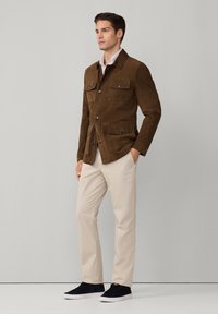 Giacca in suede marrone con quattro tasche, dettagli in bottoni e colletto, indossata sopra una camicia bianca e pantaloni beige.