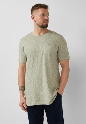 Mann mit kurzem Bart, der ein hellgrünes T-Shirt mit kurzen Ärmeln und Brusttasche sowie eine dunkle Hose trägt, seitlich steht und eine Hand in der Tasche hat.