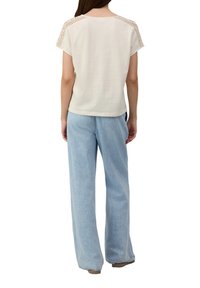 T-shirt en coton beige avec des détails en crochet sur les épaules, associé à un jean large bleu clair. Simple, coupe décontractée, texture lisse.