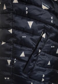 Navy puffer jacket with a quilted design featuring white triangular and circular patterns. Includes a side pocket for functionality.