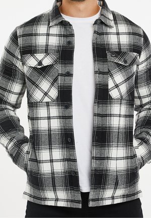 Homme portant une chemise à carreaux noir, blanc et gris boutonnée sur un t-shirt blanc uni, les mains dans les poches.