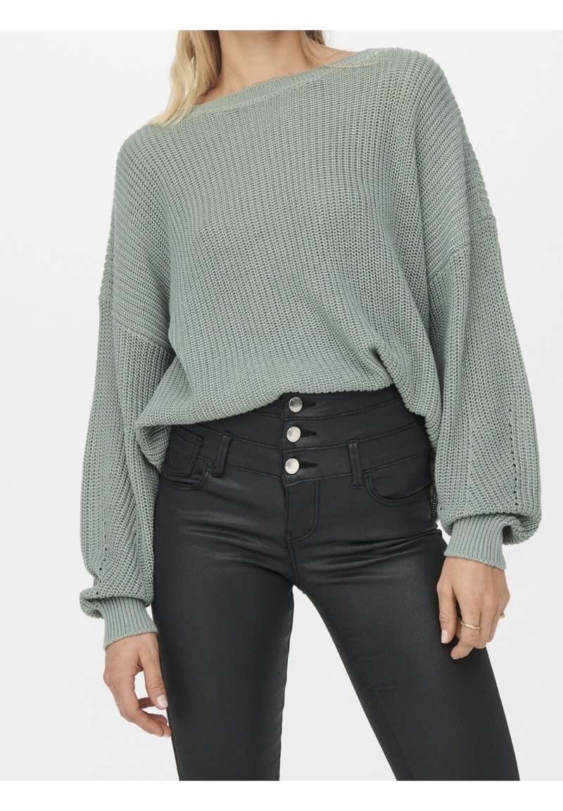 Femme portant un pull en tricot ample vert sauge et un pantalon noir taille haute avec quatre boutons argentés, debout dans une posture détendue.