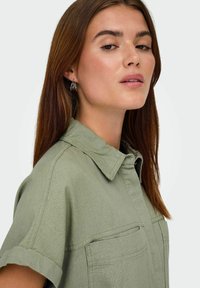 Junge Frau mit langen braunen Haaren, die silberne Creolen und ein olivgrünes Hemd mit hochgekrempelten Ärmeln trägt, vor einem schlichten Hintergrund.