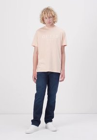 Svijetlo ružičasta pamučna majica s bijelim logotipom "GUESS EST. 1981", u kombinaciji s tamnoplavim trapericama i bijelim tenisicama. Jednostavan, ležeran outfit.